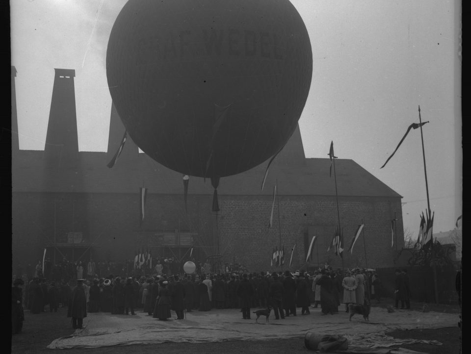 Dans quelle ville se situe cette préparation d'une ascension en ballon et quelle industrie représente ce bâtiment ?